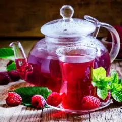 Jak zrobić herbatę malinową? Szklanka aromatycznego napoju z malinami i miętą, obok szklanego dzbanka z naparem.